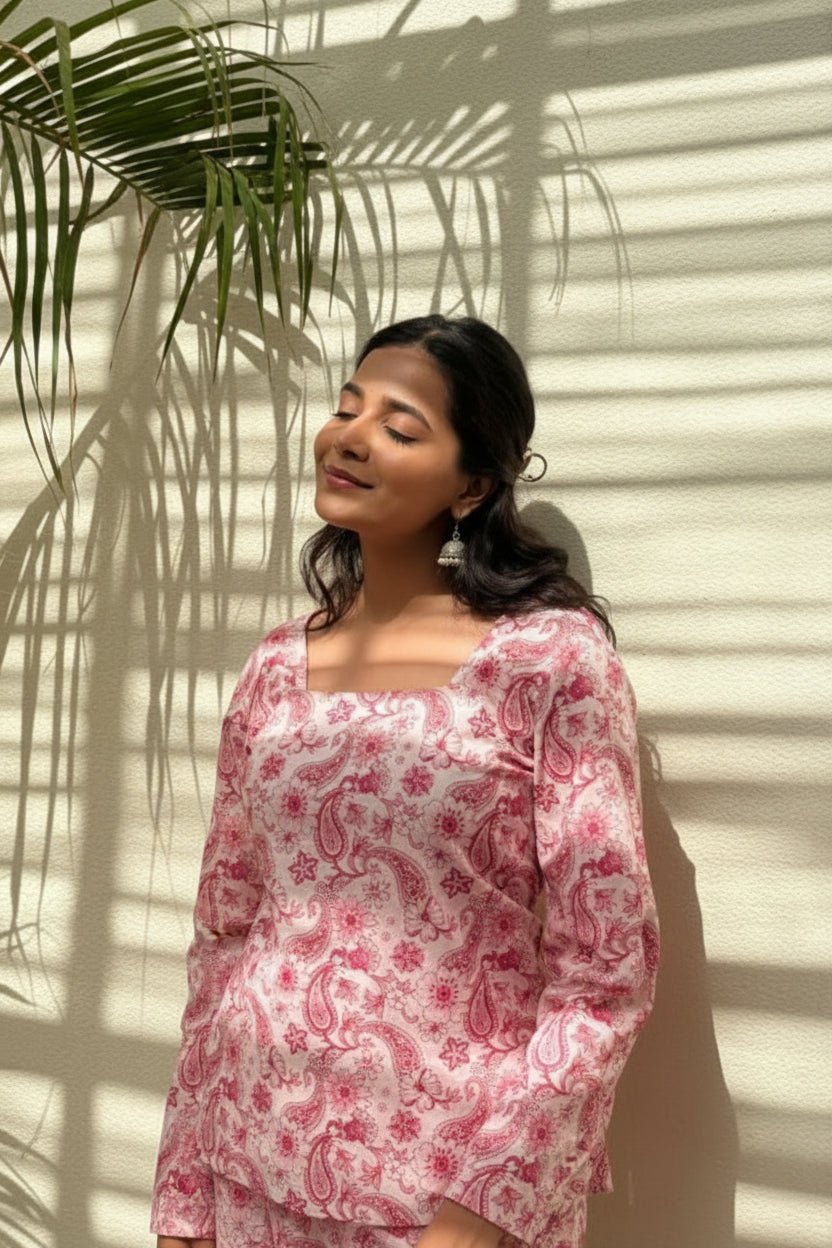 Woman wearing a pink floral blouse and white pants standing against a light-colored wall with shadows.